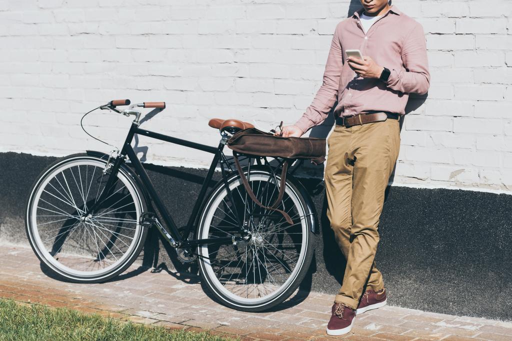 Μερική άποψη του ανθρώπου σε κομψά ρούχα χρησιμοποιώντας smartphone ενώ στέκεται κοντά ποδήλατο στον δρόμο - Φωτογραφία, εικόνα