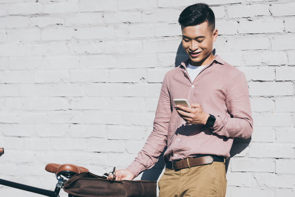 uśmiechnięty mężczyzna azjatyckich za pomocą smartfonu, stojąc w pobliżu rower na ulicy - Zdjęcie, obraz