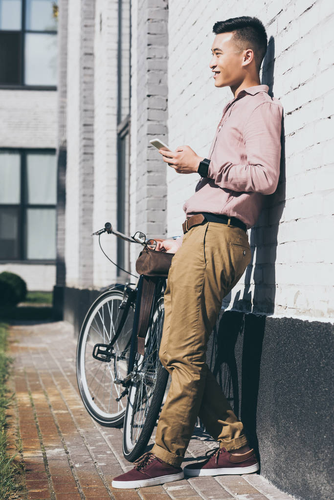 asian man in stylish clothing with smartphone standing near bicycle on street - Photo, Image