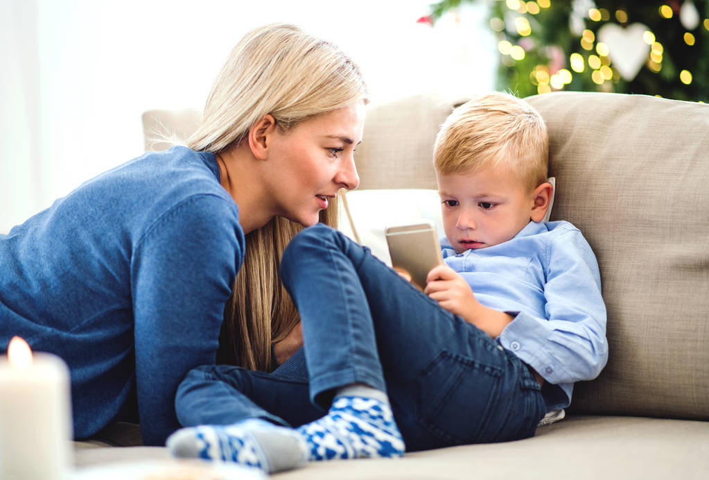 母と小さな男の子がクリスマスの時に自宅のソファの上に座ってスマート フォン. - 写真・画像