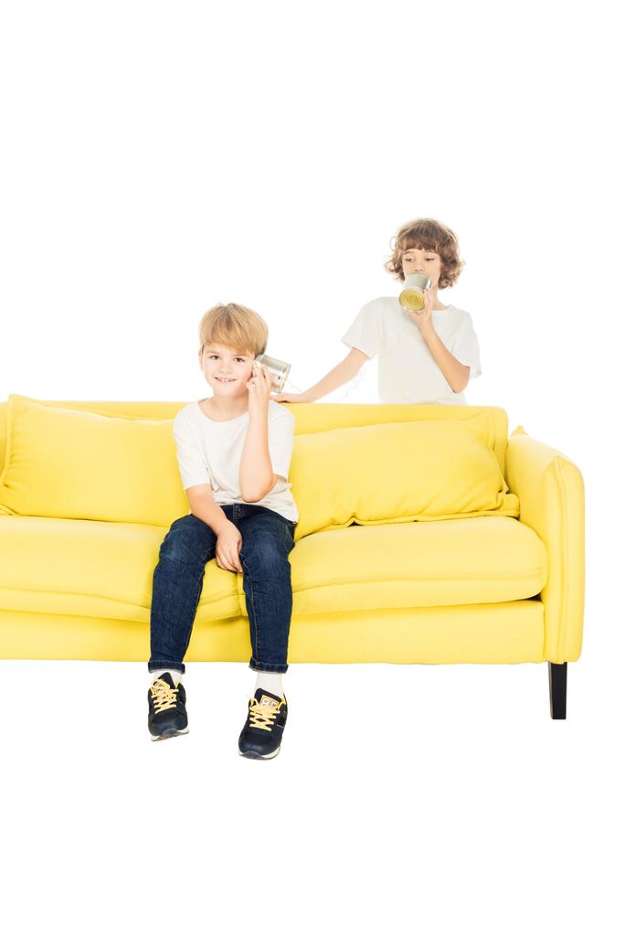 cheerful boys playing with tin cans phone on yellow sofa isolated on white - Photo, Image