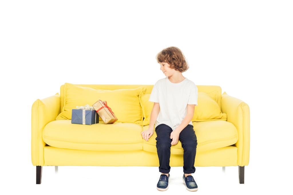 αξιολάτρευτο παιδί κάθεται στο κίτρινο καναπέ και να βλέπουν κουτιά δώρων που απομονώνονται σε λευκό - Φωτογραφία, εικόνα