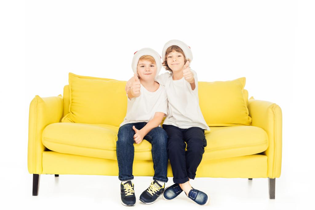 αξιολάτρευτα αγόρια καπέλα santa κάθονται στον καναπέ κίτρινο απομονωθεί σε λευκό και δείχνει τους αντίχειρες επάνω - Φωτογραφία, εικόνα