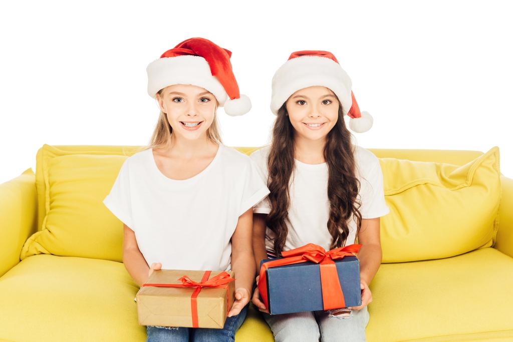 smiling adorable kids in santa hats sitting on yellow sofa with presents isolated on white - Photo, Image