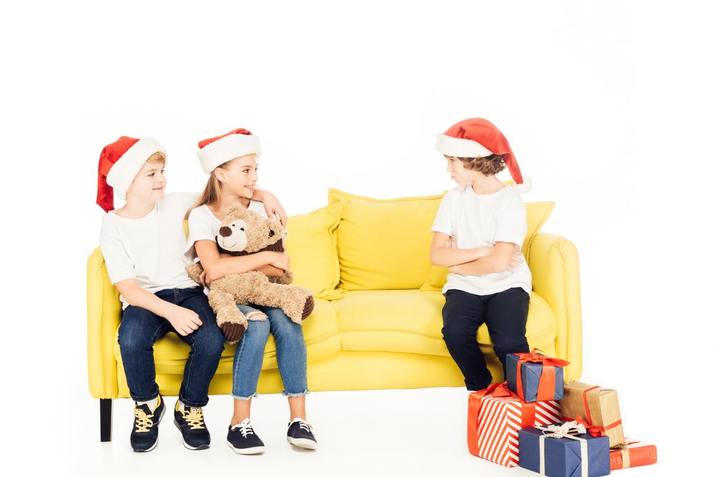 adorable kids in santa hats hugging on yellow sofa, angry boy sitting with crossed arms isolated on white - Photo, Image