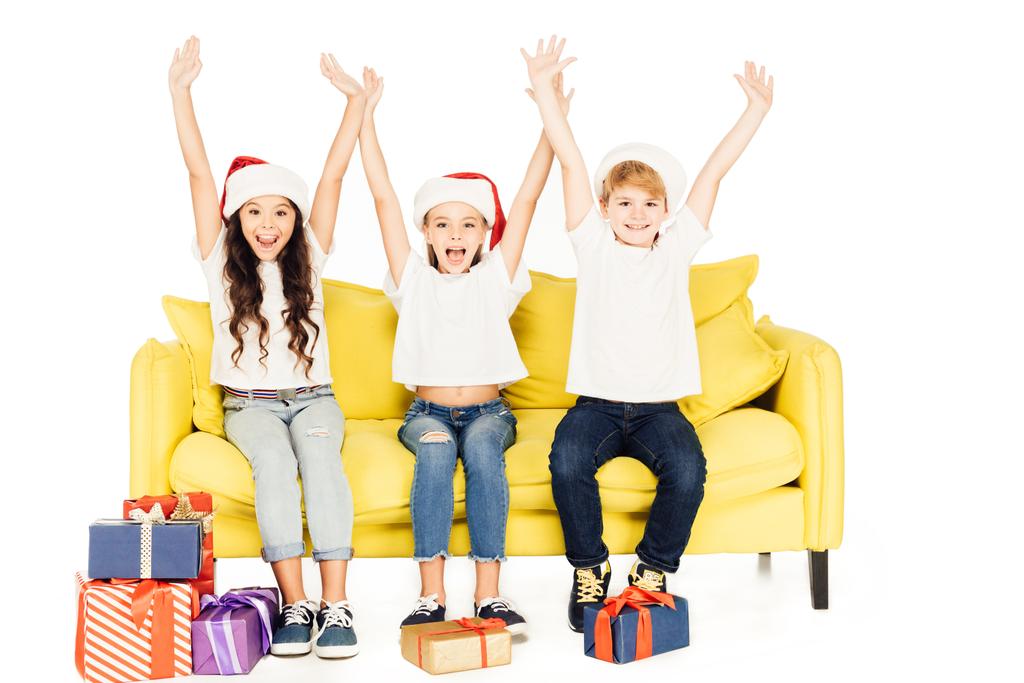 glücklich entzückende Kinder in Weihnachtsmannmützen sitzen auf dem gelben Sofa mit erhobenen Händen isoliert auf weiß - Foto, Bild