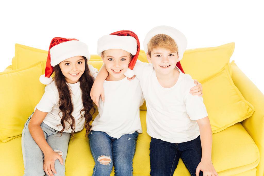 visão de alto ângulo de crianças adoráveis sorridentes em chapéus de santa sentado no sofá amarelo e olhando para a câmera isolada no branco
 - Foto, Imagem