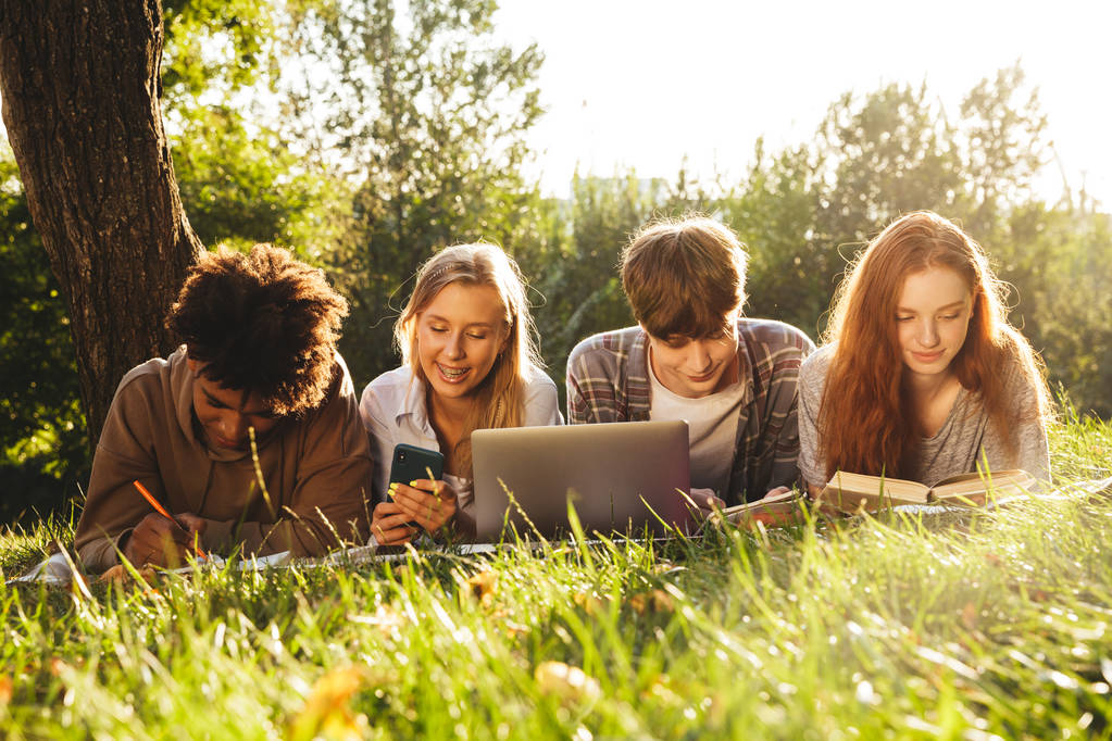 Gruppe fröhlicher multhischer Schüler, die gemeinsam im Park Hausaufgaben machen, Laptop und Handy benutzen und auf einer Wiese liegen - Foto, Bild