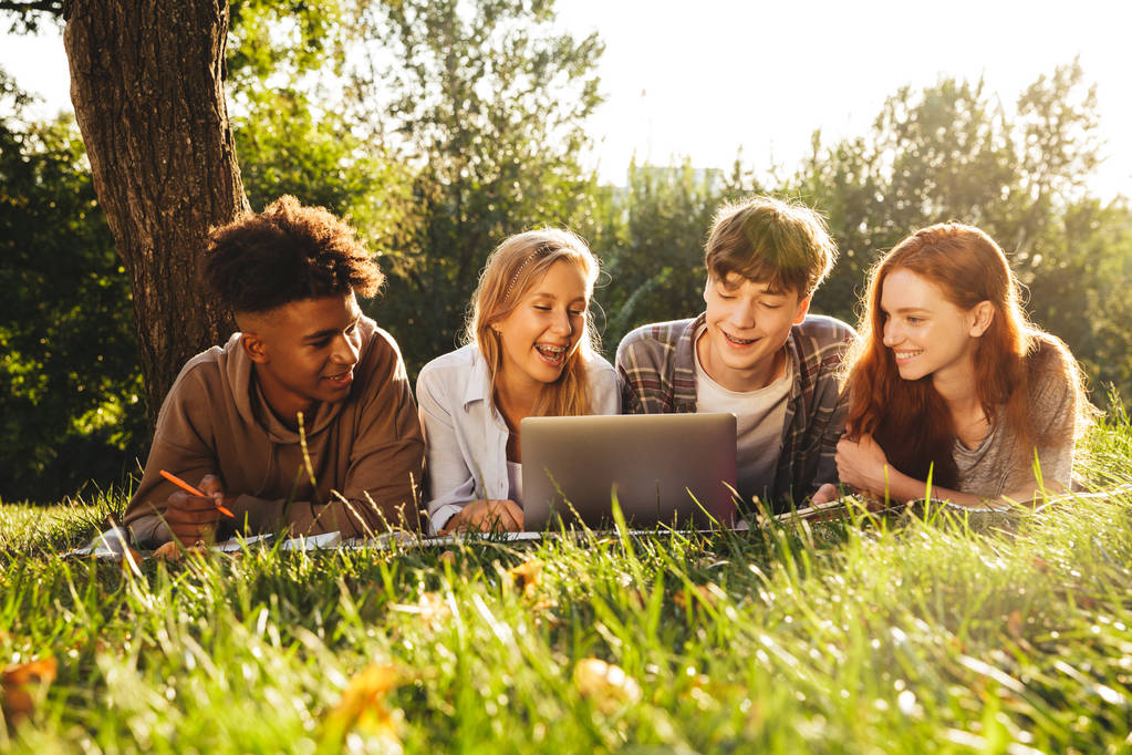 Ryhmä iloisia monikansallisia opiskelijoita tekemässä läksyjä yhdessä puistossa, käyttäen kannettavaa tietokonetta ja matkapuhelinta, makaamassa ruohikolla.
 - Valokuva, kuva