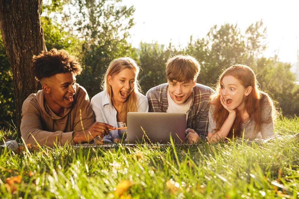 Ryhmä iloisia monikansallisia opiskelijoita tekemässä läksyjä yhdessä puistossa, käyttäen kannettavaa tietokonetta, makaamassa ruohikolla.
 - Valokuva, kuva