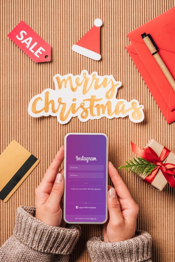 частичный просмотр женщины, держащей смартфон с Instagram на экране над поверхностью с табличкой продажи, кредитной картой и счастливым рождественским надписью
 - Фото, изображение