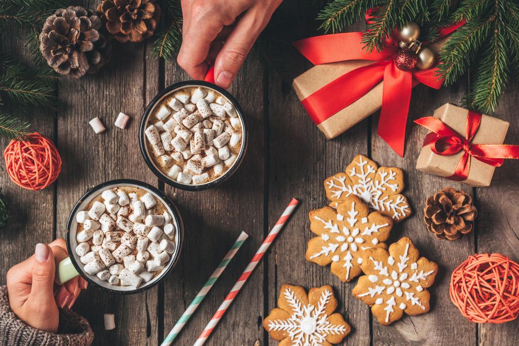 oříznutý pohled páru držícího kakaa košíčky s marshmallows a perník na dřevěné pozadí s vánoční výzdobou - Fotografie, Obrázek