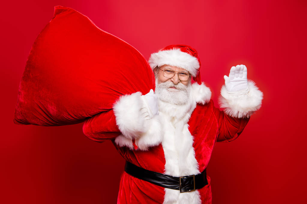 Vítejte na slavnostní akci party! Neopatrný bezstarostné dědeček stylový Santa čepice brýle bílé vousy drží tašku na rameno pohled na kamery mával, nashledanou nebo Ahoj izolované na červeném pozadí - Fotografie, Obrázek