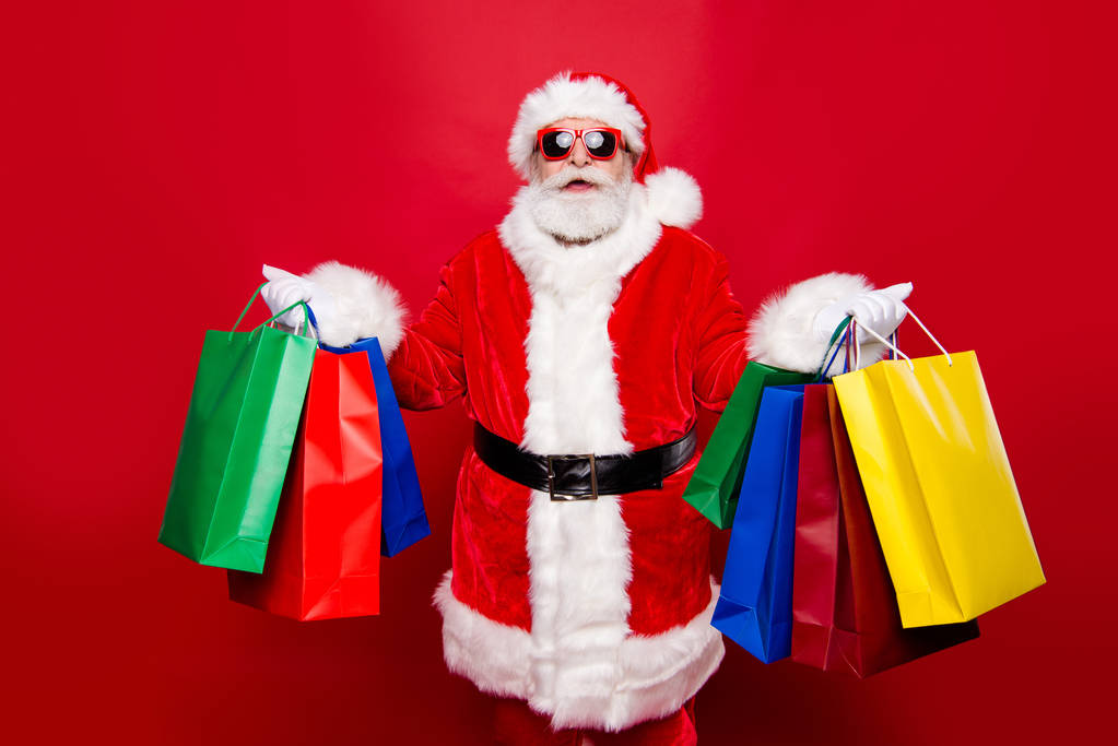 Vánoce zima noel eve ve věku zralé stylový dědeček Mikuláš v tradici čepice kostým brýle s bílým vousem držet barvu koupit tašky v rukou izolované na červeném pozadí - Fotografie, Obrázek