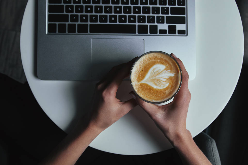 Schnappschuss von Person, die Tasse mit Cappuccino hält und Laptop benutzt - Foto, Bild
