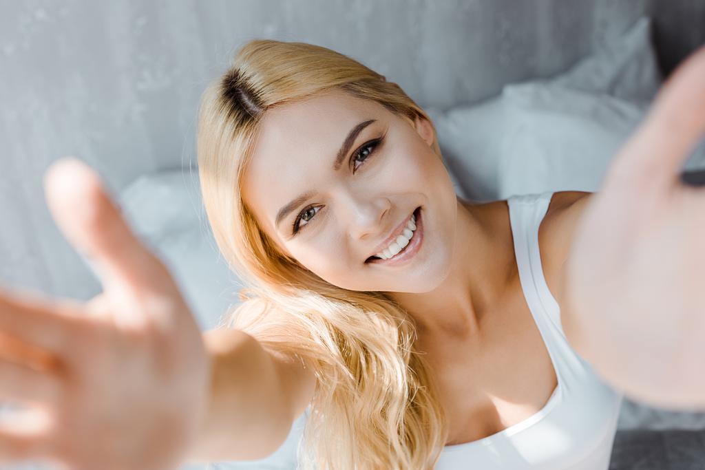 enfoque selectivo de la mujer rubia feliz alcanzando las manos y sonriendo a la cámara en el dormitorio
 - Foto, Imagen