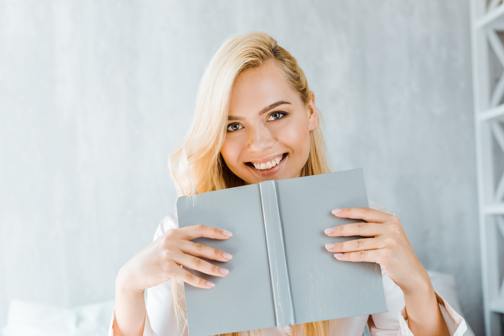 feliz joven mujer sosteniendo libro y sonriendo a la cámara en el dormitorio
 - Foto, Imagen