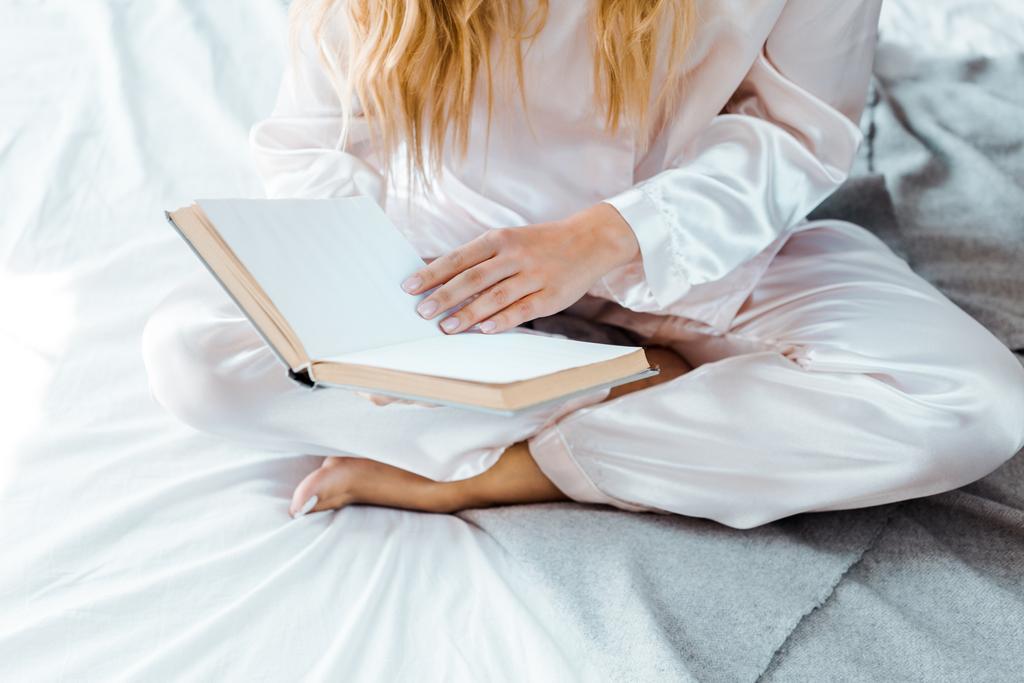 oříznuté záběr mladé ženy v pyžamu, sedí na posteli a čtení knihy - Fotografie, Obrázek