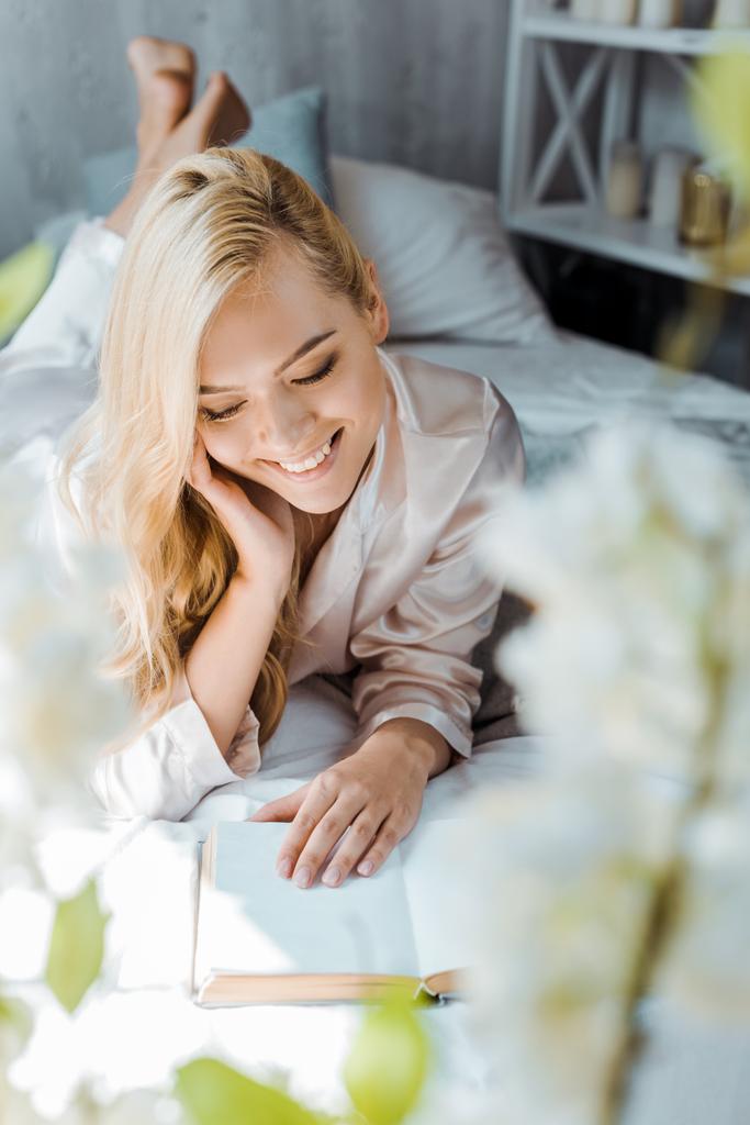 selective focus of beautiful smiling girl in pajamas lying on bed and reading book - Photo, Image