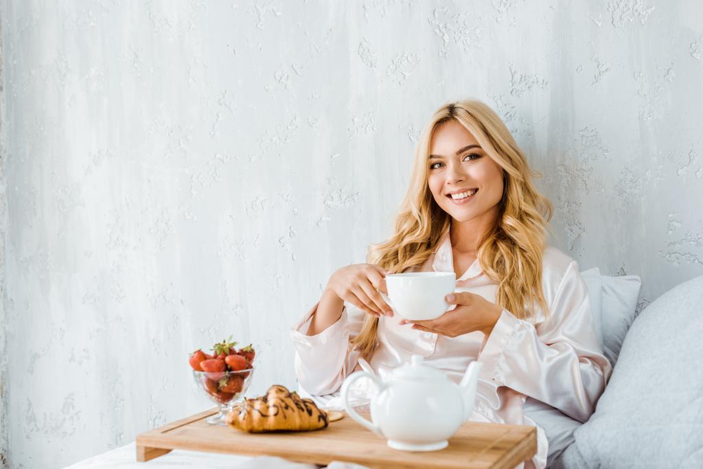 krásná usmívající se žena hospodářství šálek čaje v posteli ráno a při pohledu na fotoaparát - Fotografie, Obrázek