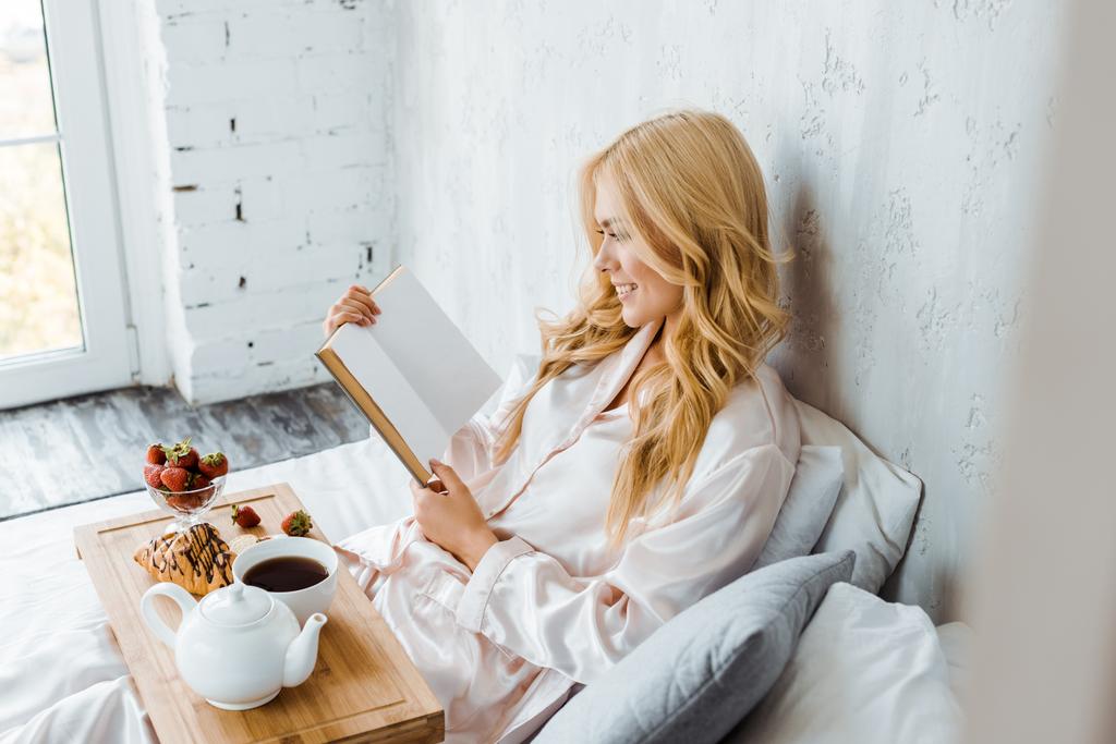 attractive woman in pajamas reading book, breakfast on wooden tray in bed - Photo, Image