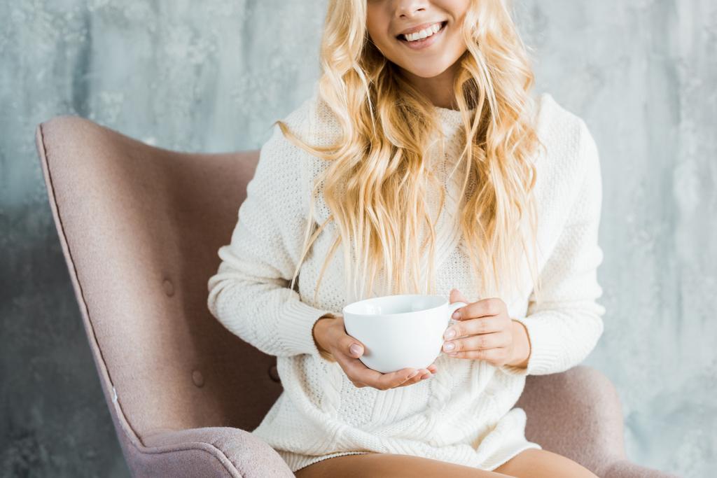 セーターの肘掛け椅子に座っていると寝室で朝の一杯のコーヒーを保持している女性のトリミングされた画像 - 写真・画像