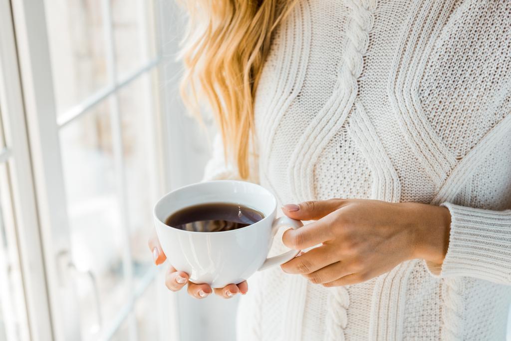 自宅の窓の近くの一杯のコーヒーを保持しているセーターの女性のトリミングされた画像 - 写真・画像
