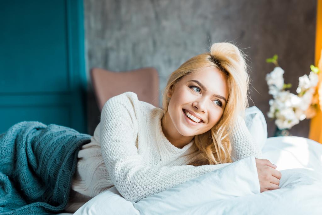 happy attractive woman in sweater lying under blanket on bed in bedroom and looking away - Photo, Image