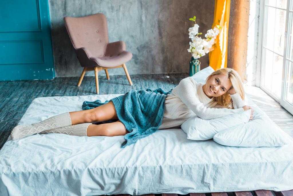 sonriente atractiva mujer en suéter acostado debajo de la manta en la cama en el dormitorio y mirando a la cámara
 - Foto, imagen