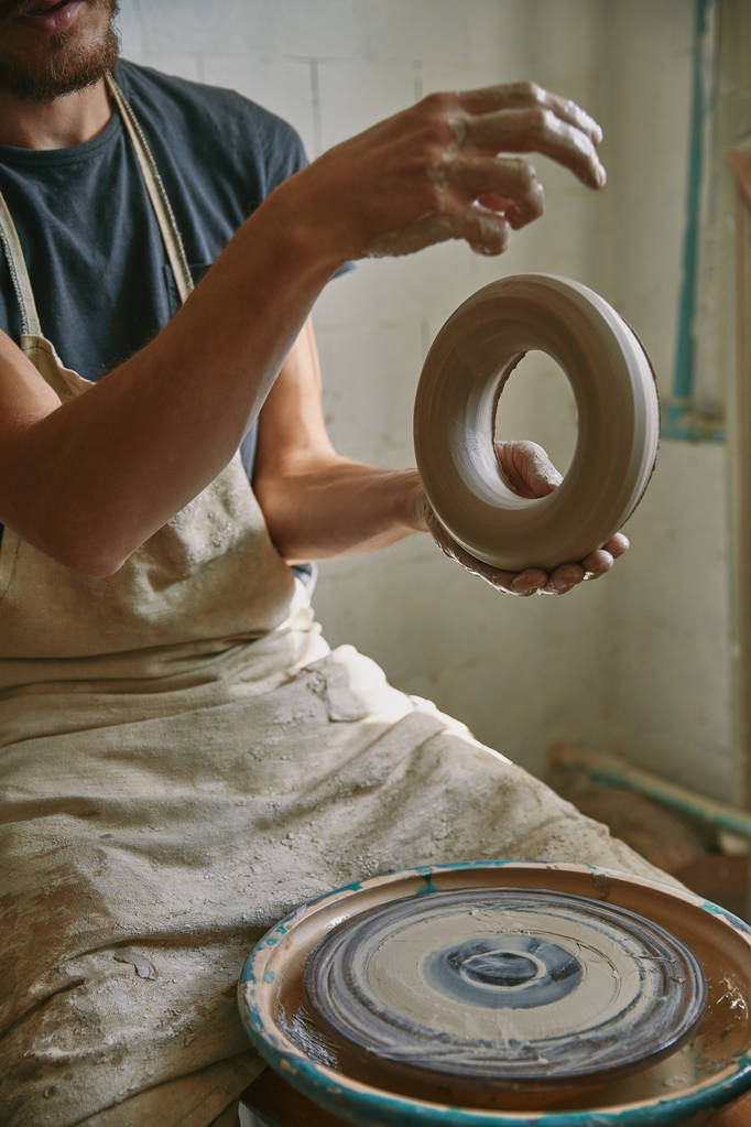 частичный вид профессионального гончара в фартуке, держащего глину в мастерской керамики
 - Фото, изображение