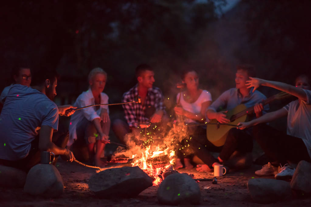 un grupo de jóvenes amigos felices relajarse y disfrutar de la noche de verano alrededor de la fogata en la orilla del río - Foto, imagen