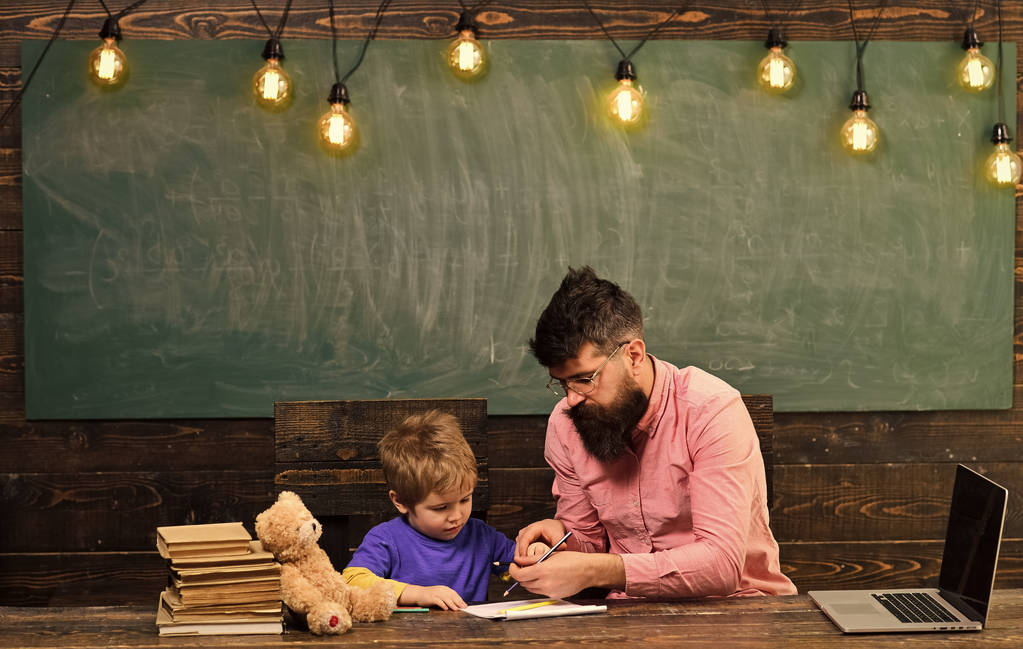 Μαθητή με δάσκαλο στο σχολείο. Δασκάλου που βοηθούν το παιδί να γράψει γράμματα στο copybook. Άνδρας και αγόρι κάθεται στο γραφείο με laptop και σωρός από βιβλία - Φωτογραφία, εικόνα