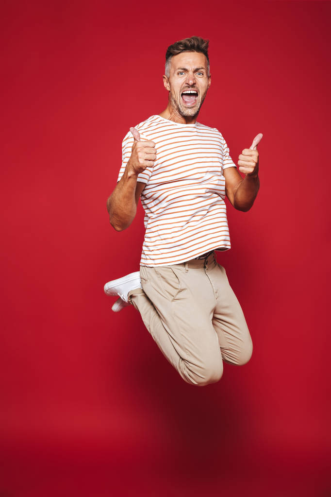 ジャンプして赤い背景に分離親指を示すストライプ t シャツで幸せな男の全身写真 - 写真・画像