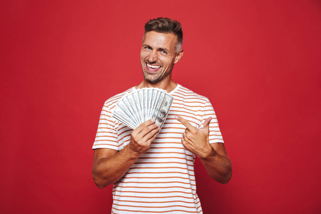 Image d'un homme non rasé en t-shirt rayé souriant et tenant fan d'argent en espèces isolé sur fond rouge
 - Photo, image