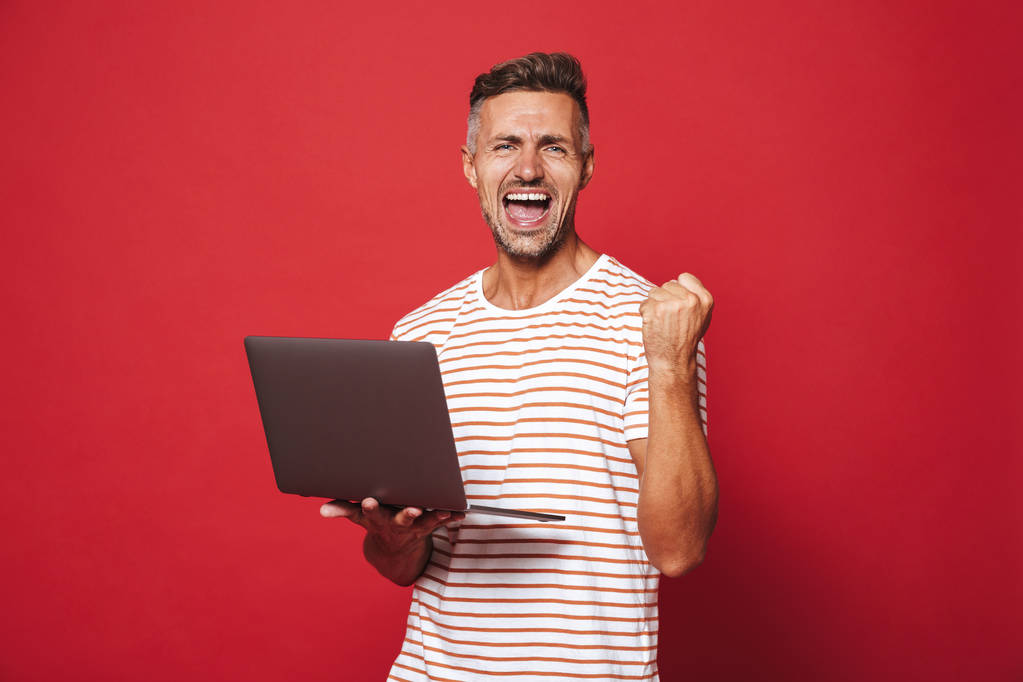Bild des erfolgreichen Typen im gestreiften T-Shirt lächelt und hält grauen Laptop isoliert über rotem Hintergrund - Foto, Bild