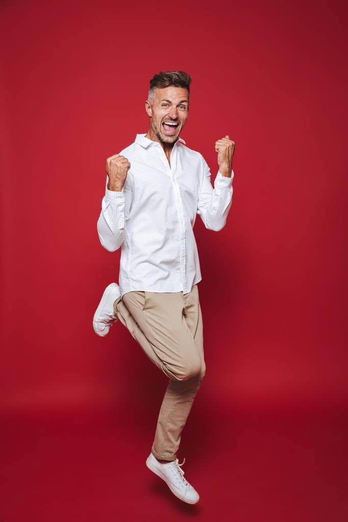 Volle lengte foto van gelukkig man in formele slijtage schreeuwen en balde vuisten geïsoleerd op rode achtergrond - Foto, afbeelding