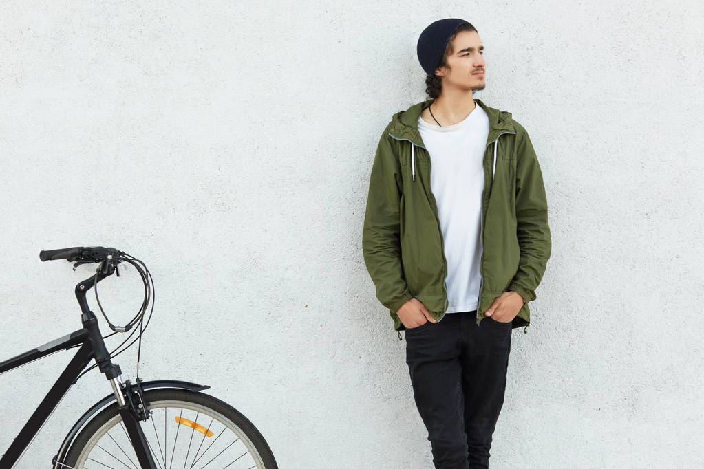 若さとサイクリングの概念。帽子、緑色のジャケットに身を包んだスタイリッシュな男、瞑想的な表情で脇に焦点を当てて、スポーツ自転車乗り場のポケットに手を続けていると思って接して自転車で旅. - 写真・画像