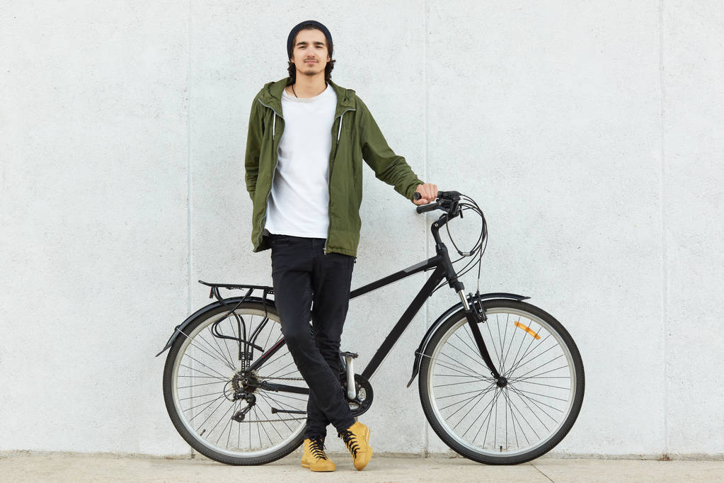 Ізольований знімок стильного підлітка-хіпстера, одягненого в стильний одяг і кросівки, стоїть біля спортивного велосипеда, насолоджується відпочинком один, рекламує свій велосипед, позує над білою бетонною стіною
. - Фото, зображення