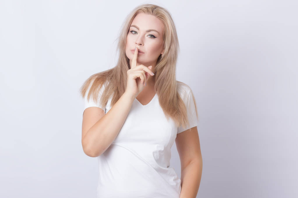 Bild einer schönen blonden Frau, die den Finger in der Nähe ihrer Lippen hält. isoliert über grauem Hintergrund - Foto, Bild