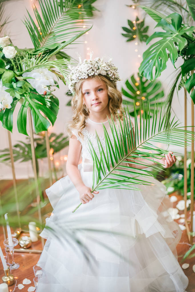 Menina bonito com cabelo encaracolado loiro em um vestido de noiva branco e uma grinalda de flores em decorações florais feitas de folhas tropicais, rosas brancas e orquídeas
 - Foto, Imagem