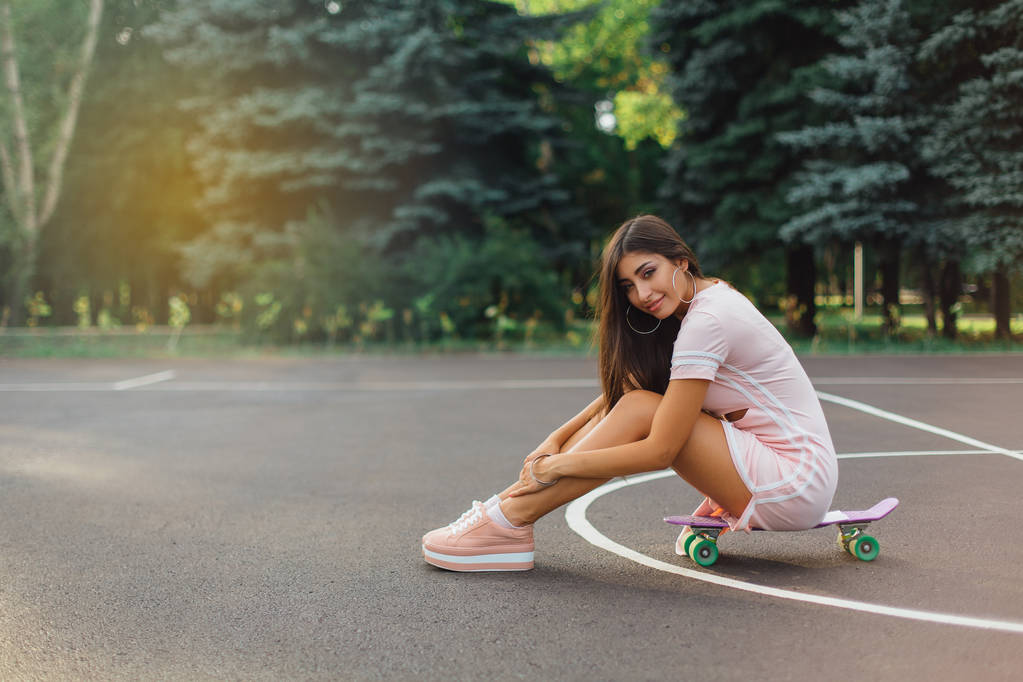 Portrait d'une charmante brune souriante assise sur son skateboard sur un terrain de basket. Femme heureuse avec look tendance prenant une pause pendant le coucher du soleil. - Photo, image
