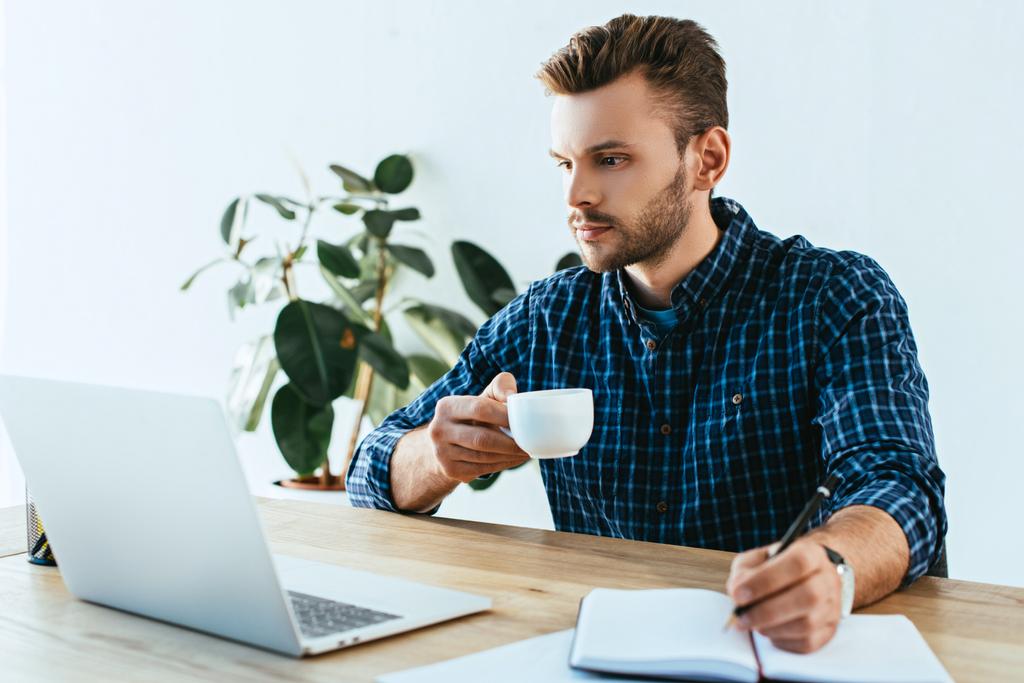 joven hombre de negocios con taza de café que participa en webinar en la mesa con ordenador portátil en la oficina
 - Foto, Imagen