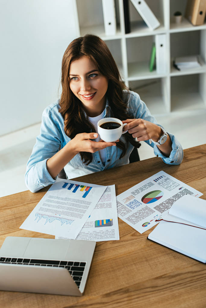 joven empresaria con taza de café en el lugar de trabajo con papeles y portátil en la oficina
 - Foto, imagen