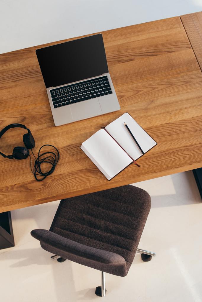 vista dall'alto di laptop con schermo bianco, cuffie e notebook su tavolo in legno con sedia da ufficio nelle vicinanze
 - Foto, immagini