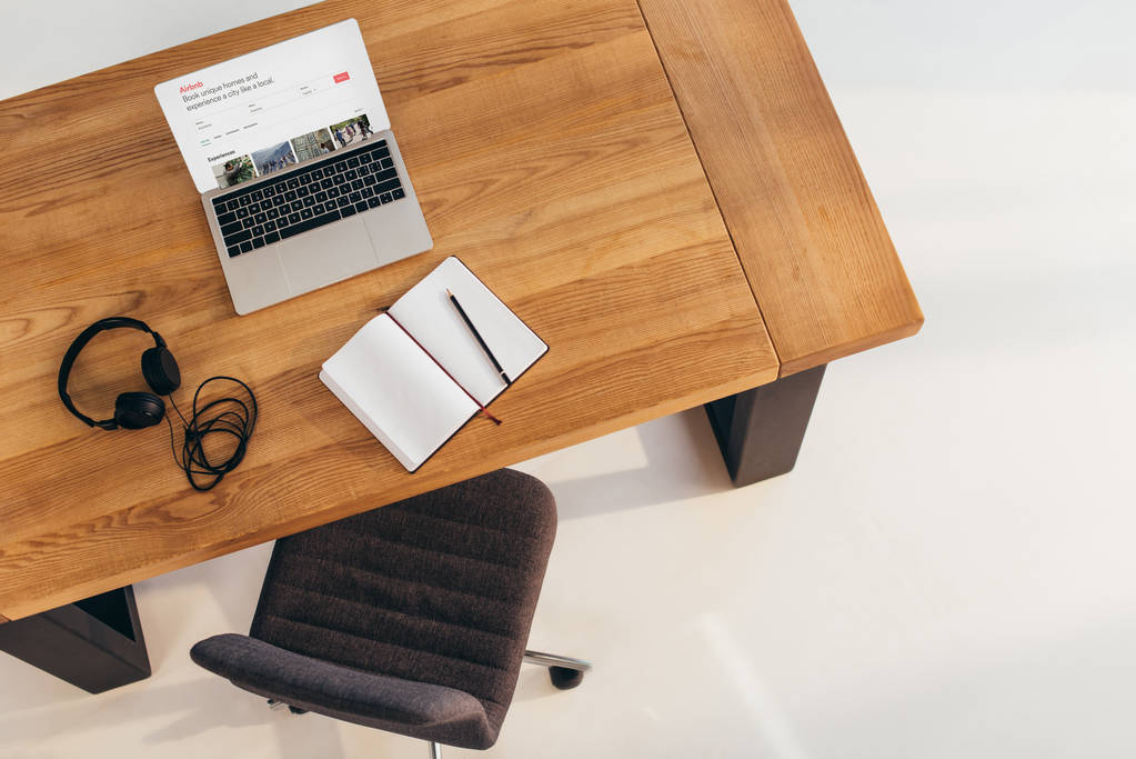 Draufsicht auf Laptop mit Airbnb-Webseite, Kopfhörer und Notizbuch auf Holztisch mit Bürostuhl in der Nähe - Foto, Bild