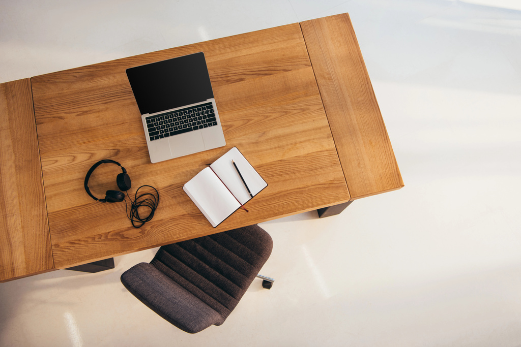 top view of laptop with blank screen, headphones and notebook on wooden table with office chair near by - Photo, Image