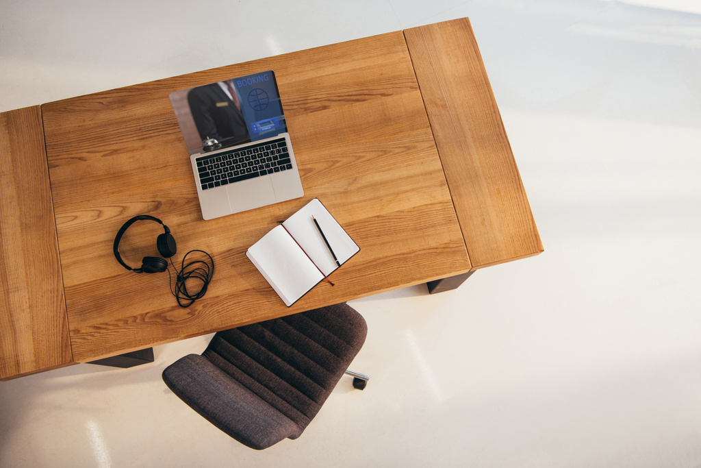 Widok z góry laptopa z napisem rezerwacji na ekranie słuchawki i notebook na drewnianym stole z krzesłem biurowym w pobliżu przez - Zdjęcie, obraz