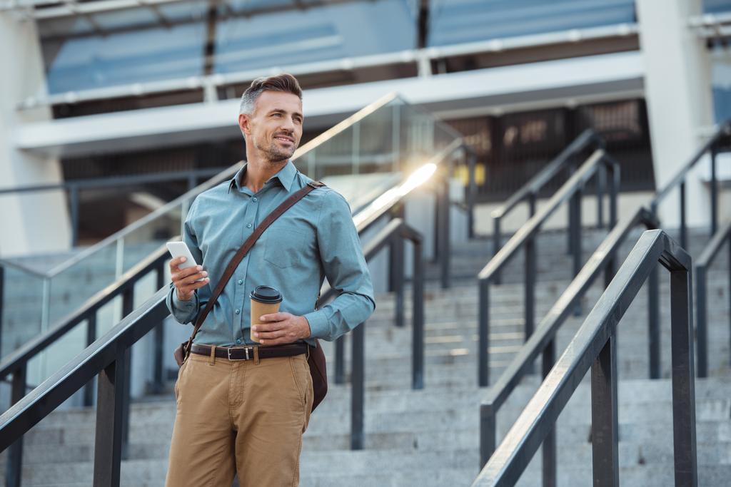 sonriente hombre de negocios de mediana edad con café para llevar y smartphone mirando hacia otro lado en las escaleras
 - Foto, Imagen