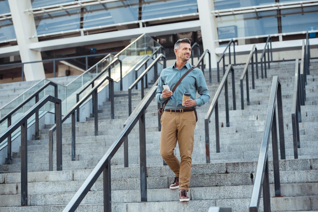 улыбающийся бизнесмен средних лет, держащий бумажную чашку и смартфон, спускаясь по лестнице
 - Фото, изображение