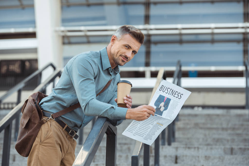 knappe man holding bedrijf krant en koffie te gaan terwijl staande op de trap en lachend op camera - Foto, afbeelding